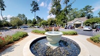 Located at 8439 Dorchester Rd in North Charleston, South Carolina, this single-story property makes a strong first impression with its thoughtfully designed exterior and inviting landscaping. At the heart of the entrance area stands a decorative tiered fountain set within a circular basin lined with smooth river stones, creating a serene and polished focal point for visitors arriving at the property. Surrounding the fountain, manicured shrubbery, blooming rose bushes, and mature trees — including towering Southern pines — frame the grounds with natural beauty and shade. The landscaping is well-maintained, with mulched beds and neatly trimmed hedges that contribute to a clean, professional aesthetic. A paved roundabout driveway provides convenient access and ample parking. The building itself is a modest, light-colored structure with a classic architectural style that complements the natural surroundings. The overall ambiance of the property is calm and welcoming, blending functional commercial design with attractive outdoor elements. The combination of the water feature, established trees, and manicured greenery creates an environment that feels both professional and approachable, reflecting a space that prioritizes curb appeal and a positive visitor experience from the moment of arrival.