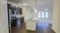 This single-floor residence offers a thoughtfully designed open-concept layout that seamlessly connects the kitchen and living area. The kitchen is a standout feature, showcasing dark espresso cabinetry that contrasts elegantly with light granite countertops. Stainless steel appliances, including a range and over-the-range microwave, add a modern, polished touch. A central kitchen island with a built-in sink and matching granite surface provides additional prep space and serves as a natural gathering point. Track lighting above the kitchen and stylish pendant lights over the island ensure excellent task illumination throughout. Rich, dark hardwood flooring extends across the main living area, lending warmth and sophistication to the space. Crown molding along the ceilings adds an architectural detail that elevates the overall aesthetic. The adjoining living area transitions to carpet underfoot and benefits from abundant natural light streaming through a set of large windows and French doors, which also offer views to the outdoors. A ceiling fan in the living area enhances comfort year-round. The overall ambiance is clean, contemporary, and inviting, making this space well-suited for both everyday living and entertaining.