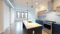 This single-floor unit showcases an industrial-modern aesthetic rooted in loft-style design. Polished concrete floors extend throughout the space, complementing exposed ductwork on the ceilings and adding to the raw, urban character. Large black-framed windows are present in multiple rooms, flooding the living areas with natural light and offering elevated views of the surrounding cityscape.

The open-concept kitchen serves as a central feature of the living area, anchored by a generous island with a quartz waterfall-style countertop and an integrated sink with a professional-style faucet. Navy blue lower cabinetry is paired with white subway tile, open natural wood shelving, a stainless steel range with gas burners, a wall-mounted range hood, and a full-size stainless refrigerator.

The living area is spacious and bright, with whitewashed brick walls adding textural depth. A separate bedroom offers city views through black-framed windows, while the bathroom features dark penny-round tile flooring, a frameless glass walk-in shower, and a quartz vanity with a wood-toned cabinet. A dedicated laundry closet houses a stacked washer and dryer unit for added convenience.