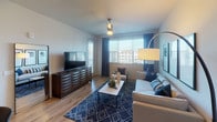 This thoughtfully designed single-floor residence offers a seamless blend of modern style and functional living. The open-concept kitchen is a standout feature, equipped with stainless steel appliances including a French door refrigerator, electric range, and microwave. Crisp white cabinetry extends to the ceiling, complemented by a light quartz countertop island with bar seating and elegant pendant lighting overhead. Wood-look flooring flows throughout the main living areas, connecting the kitchen to a bright living room that benefits from generous natural light through plantation-shuttered windows. The living area provides ample space for comfortable seating and entertainment. The residence includes multiple bedrooms, each carpeted for comfort and fitted with ceiling fans, large windows with natural light, and room for substantial furniture. The primary bedroom is served by a sleek en-suite bathroom featuring a floating vanity, large-format tile flooring, and direct access to a walk-in closet. A second full bathroom mirrors the same clean, contemporary aesthetic with white cabinetry and tiled floors. Throughout the home, a neutral color palette and recessed lighting create a calm, cohesive ambiance well-suited for modern living.