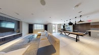 This single-floor amenity space is thoughtfully designed with a contemporary aesthetic, combining multiple functional zones within an open, cohesive layout finished in neutral tones, large-format tile flooring, and recessed lighting throughout.

The lounge and media area anchors one end of the space, featuring a generously sized sectional sofa, an accent armchair, and a glass coffee table arranged on a textured area rug. A large wall-mounted flat-screen television is set against a dark wood-paneled feature wall, complemented by framed abstract artwork, creating a polished, relaxed atmosphere.

The open central area transitions seamlessly into a game and dining zone, where a long communal game table sits beneath a cluster of striking black pendant lights. Adjacent seating with high-top stools lines the perimeter near large windows, allowing natural light to fill the space and providing outdoor views.

The kitchen area is equipped with a central island featuring bar-height seating for multiple guests, warm wood cabinetry, a mosaic tile backsplash, built-in televisions, and ample counter space — well-suited for catering or social gatherings.

The dining nook offers several café-style tables and chairs beside floor-to-ceiling windows, brightened by natural light and accented with colorful artwork, completing this versatile and well-appointed communal space.