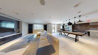 This single-floor amenity space is thoughtfully designed with a contemporary aesthetic, combining multiple functional zones within an open, cohesive layout finished in neutral tones, large-format tile flooring, and recessed lighting throughout.

The lounge and media area anchors one end of the space, featuring a generously sized sectional sofa, an accent armchair, and a glass coffee table arranged on a textured area rug. A large wall-mounted flat-screen television is set against a dark wood-paneled feature wall, complemented by framed abstract artwork, creating a polished, relaxed atmosphere.

The open central area transitions seamlessly into a game and dining zone, where a long communal game table sits beneath a cluster of striking black pendant lights. Adjacent seating with high-top stools lines the perimeter near large windows, allowing natural light to fill the space and providing outdoor views.

The kitchen area is equipped with a central island featuring bar-height seating for multiple guests, warm wood cabinetry, a mosaic tile backsplash, built-in televisions, and ample counter space — well-suited for catering or social gatherings.

The dining nook offers several café-style tables and chairs beside floor-to-ceiling windows, brightened by natural light and accented with colorful artwork, completing this versatile and well-appointed communal space.