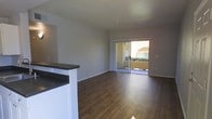 This single-story space features an open-concept layout that seamlessly connects the kitchen and living areas. The flooring throughout is a rich, dark hardwood that adds warmth and contrast against the neutral gray-white walls and white baseboards. A kitchen counter with a dark surface is partially visible, forming a natural divider between the cooking and living zones while maintaining the open feel of the space.

The living area is generously sized, offering flexible arrangement possibilities. A large sliding glass door dominates the far wall, functioning as both a primary light source and access point to an outdoor area. Through the glass, greenery and the exterior of an adjacent building are visible, providing a pleasant outdoor view. The abundant natural light that flows through the sliding door brightens the interior, enhancing the airy, open ambiance of the space.

The walls are smooth and painted in a clean, neutral tone, complementing the overall minimalist aesthetic. The space is move-in ready, with no obstructions, making it easy to visualize furniture placement and personalization. The single-floor layout ensures convenient accessibility throughout.