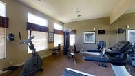 This fitness center offers a practical and well-organized space designed to support a full range of workout routines. The room is finished with neutral, warm-toned walls and carpeted flooring, creating a comfortable environment for exercise. Large windows fitted with blinds and decorative valances line one wall, allowing natural light to fill the space while maintaining privacy. The facility is equipped with a multi-station cable strength machine, providing users with a wide variety of resistance training options. Cardio equipment includes an elliptical trainer and at least one stationary bike, giving members multiple options for cardiovascular workouts. A wall-mounted flat-screen television is positioned for easy viewing during exercise sessions. Framed artwork on the walls adds a finishing touch to the space, contributing to an inviting atmosphere. The ceiling features a ventilation system to ensure proper air circulation throughout workouts. The single-floor layout keeps everything accessible and easy to navigate, making this fitness center a convenient and fully functional amenity for regular use.