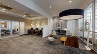 This thoughtfully designed single-floor amenity space offers a diverse range of gathering areas unified by a contemporary aesthetic. The main lounge area is anchored by a large sectional sofa, warm leather armchairs, and a live-edge wood coffee table arranged beneath a series of bold pendant lights. A gallery wall of vibrant blue abstract artwork adds a curated artistic touch, while dark wood-toned flooring runs throughout, grounding the overall design.

Adjacent to the lounge, a billiards table serves as a focal point for recreation, positioned near a wet bar with wood-paneled cabinetry and bar stool seating. A dining area with an oval dark stone table and upholstered chairs provides a more formal gathering spot, positioned close to a sun-drenched nook enclosed by floor-to-ceiling windows that offer lush outdoor views and abundant natural light. High-top tables and modern bucket chairs furnish this bright corner.

The full kitchen is outfitted with dark espresso cabinetry, granite countertops, a stone tile backsplash, and a complete suite of stainless steel appliances, making it well-equipped for catered events or communal use. Throughout, recessed lighting, pendant fixtures, and clean architectural lines contribute to a polished, welcoming atmosphere.