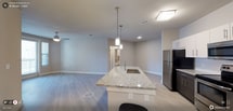 This two-bedroom, two-bathroom residence is thoughtfully designed on a single floor, offering a cohesive and functional layout. The open-concept living and kitchen area serves as the heart of the home, featuring light-toned luxury vinyl plank flooring that extends throughout, creating a clean and contemporary aesthetic.

The kitchen is a standout feature, centered around a large island with a striking granite countertop and an integrated sink. Pendant lighting hangs above the island, providing both task lighting and a stylish focal point. A stainless steel refrigerator is visible nearby, complementing the modern finishes. Recessed lighting and flush-mount fixtures illuminate the space efficiently.

The open layout connects the kitchen seamlessly to the living area, which offers ample room for furnishings and everyday use. Neutral gray walls and white trim provide a versatile backdrop throughout. Hallways lead to the two bedrooms and bathrooms, with a glimpse of a well-appointed bathroom visible in the background. Ceiling vents indicate a central HVAC system for year-round comfort. Overall, the space presents a bright, move-in-ready environment with clean, modern details.