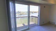 This single-floor residence offers a well-organized layout designed for comfortable everyday living. The galley-style kitchen is efficiently arranged with rich wood cabinetry, granite-look countertops, a double stainless steel sink, and white appliances including a gas range and full-size refrigerator. A hardwood-style floor runs through the kitchen, which opens directly into the adjoining dining or living area, illuminated by a classic chandelier overhead. Natural light flows through a large window at the far end, creating a bright and welcoming atmosphere. The living area features plush carpeting and leads to a set of sliding glass doors that open onto a private wooden balcony. This outdoor space is a standout feature of the home, offering sweeping views of a serene lake framed by lush, tree-lined shores — an ideal setting for relaxing or enjoying seasonal scenery. The overall ambiance of the space is cozy and practical, with a neutral color palette throughout that provides a versatile backdrop for a variety of interior styles. The connection between indoor living and the tranquil waterfront view gives this residence a distinctive and appealing character.
