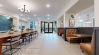 Located at 9821 Research Drive in Charlotte, NC, this single-floor amenity space offers a well-appointed and versatile communal environment designed for socializing, relaxation, and productivity. The layout is open and generously proportioned, with smooth concrete-style flooring, crisp white walls, and a consistent palette of warm browns, deep charcoals, and natural wood tones throughout.

The main lounge area is a standout feature, anchored by a large-screen TV mounted within an ornate decorative lattice panel wall. Built-in shelving with curated books and artwork lines the dark accent wall, while a cognac leather sofa, tufted accent chairs, and a round cocktail table create an inviting seating arrangement. Adjacent to the lounge, tufted leather booth seating and wall sconces offer a more intimate setting, accented by framed animal sketch artwork.

A formal dining area accommodates a long wooden table with upholstered chairs, bathed in natural light from large windows. Black-framed glass double doors provide a seamless connection to an outdoor patio space. A game room corner features a foosball table and a pool table, with natural light streaming in through windows overlooking the surrounding urban landscape. A co-working nook with a wall-mounted white countertop and modern bar stools completes the space, offering convenient charging stations for focused work.