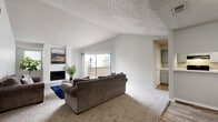 This well-appointed 2-bedroom, 3-bathroom single-floor residence offers a comfortable and functional layout across a generously sized floor plan. The open living room is a standout feature, boasting soaring vaulted ceilings with a textured finish, plush wall-to-wall carpeting, and a classic white brick fireplace that anchors the space. A sliding glass door floods the room with natural light and opens directly to a private balcony, offering pleasant outdoor views. The adjacent kitchen features a pass-through bar connecting seamlessly to the living area, complemented by gray cabinetry, light countertops, and wood-look flooring that extends through the dining and kitchen zones. Built-in shelving adds additional storage and display space near the kitchen entrance. The spacious primary bedroom includes a ceiling fan, soft carpeting, and direct access to an en-suite bathroom with gray vanity cabinetry and a tub-shower combination. A dedicated walk-in closet provides ample storage with multiple shelving rods and built-in shelving units. The second bedroom is equally well-sized, featuring corner windows that invite natural light and its own ceiling fan. Three full bathrooms ensure convenience for residents and guests alike, rounding out a practical and inviting floor plan.