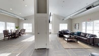 This single-floor space features a bright, open layout accentuated by soaring vaulted ceilings that create an airy, expansive atmosphere. Exposed silver ductwork runs along the upper walls, adding an industrial design element that contrasts tastefully with the otherwise clean, contemporary aesthetic. The flooring consists of patterned carpet tiles in a grey and gold tone, lending visual interest throughout the open floor plan.

The main lounge area is anchored by a dark leather sofa and loveseat arrangement set atop a neutral area rug, accompanied by a wooden coffee table. A wall-mounted flat-screen television is positioned on a structural column, serving as a central focal point for the seating area. Natural light filters generously through multiple large windows along the perimeter walls, brightening the neutral grey and white color palette.

A secondary seating area is visible toward the rear of the space, suggesting flexible use for various group sizes. Recessed lighting supplements the natural light overhead. The overall ambiance is modern and welcoming, well-suited for communal use.