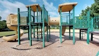 This outdoor playground is centered around a substantial multi-tower play structure finished in teal green and tan, designed to encourage active, imaginative play. Two elevated platform towers are connected by a cylindrical tunnel bridge, creating an engaging layout that promotes exploration and movement. Each tower is enclosed by vertical railing panels for safety and is topped with a tan canopy roof that provides partial shade during use.

Multiple play elements are integrated throughout the structure, including a climbing ladder with horizontal rungs offering an accessible route to the upper platforms, and at least one enclosed tube slide visible to the side of the main structure. The towers are elevated on sturdy teal steel posts, giving the play area a sense of vertical scale appropriate for school-age children.

The ground surface beneath and around the structure is covered in wood chip mulch, serving as a standard impact-absorbing safety surface. The surrounding area appears open and spacious, with a paved path and trees visible in the background under a bright, partly cloudy sky, contributing to a welcoming, park-like atmosphere.