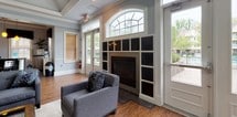This single-floor communal space is thoughtfully designed to offer a comfortable and functional gathering environment. The main lounge area is an inviting, open room anchored by a classic fireplace framed by built-in shelving and a striking arched transom window above. Warm hardwood flooring runs throughout, complementing the soft blue-gray walls and crisp white crown molding and wainscoting. A tray ceiling with recessed lighting adds architectural interest and a sense of height to the space. Large double French doors and expansive windows flood the interior with natural light while offering views of the lush greenery outside. Comfortable gray upholstered seating is arranged around a glass-top coffee table, creating a relaxed living area ideal for socializing. Adjacent to the lounge is a dedicated dining area, furnished with a dark wood dining table and cross-back chairs, accented by colorful framed artwork on the walls. A compact kitchenette rounds out the space, featuring light maple cabinetry, dark granite countertops, a stainless steel sink, and bold black tile backsplash, providing convenient amenities for gatherings. Together, these areas create a versatile, well-appointed space suited for both casual relaxation and organized events.