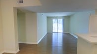 This single-story space features an open-concept living area that offers a clean, functional layout. The room is finished with rich hardwood flooring that runs throughout, adding warmth and continuity to the space. Walls are painted in a soft, neutral green tone, creating a calm and welcoming ambiance that complements the natural light flowing in from a large sliding glass door at the far end of the room. The sliding door provides direct access to an outdoor area and serves as a prominent architectural feature, flooding the interior with daylight. Recessed lighting in the ceiling supplements natural illumination, ensuring the space remains well-lit. Crisp white baseboards provide a clean contrast against both the walls and flooring. HVAC vents are integrated into the ceiling, indicating a central air system. The layout is open and unobstructed, offering flexibility for a variety of furniture arrangements. A partial wall section adds subtle architectural definition without compromising the open feel of the room. Overall, the space presents a straightforward, move-in-ready interior with practical finishes and a bright, airy atmosphere.
