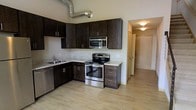 This two-floor space showcases a well-appointed kitchen that serves as a central feature of the layout. The kitchen is outfitted with rich, dark espresso-toned cabinetry that extends to the upper walls, providing generous storage capacity. Light-toned countertops with subtle veining offer a clean contrast against the darker cabinets, creating a balanced visual aesthetic. Stainless steel appliances, including a dishwasher, electric range, and over-the-range microwave, lend the kitchen a contemporary, functional character. An exposed metallic ventilation duct runs overhead, adding an industrial design element that contributes to the space's modern aesthetic. The light wood-style flooring flows seamlessly from the kitchen into the adjoining areas, enhancing the sense of continuity throughout the unit. The open layout connects the kitchen to a hallway, which leads to additional areas of the home, including access to the second floor. Neutral wall tones keep the interior feeling bright and open, while recessed and flush-mount ceiling lighting provides consistent illumination. The overall design balances modern finishes with practical functionality across both levels.