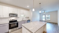 Located at 1300 S Davis Rd in LaGrange, Georgia, this well-appointed single-story home offers four bedrooms and two bathrooms across a comfortable and efficiently designed floor plan. The kitchen serves as a central highlight, boasting a large granite-topped island with pendant lighting overhead, white upper cabinetry, gray lower cabinets, a classic subway tile backsplash, and a full suite of stainless steel appliances including a French door refrigerator, electric range, microwave, and dishwasher. Light wood-look flooring runs throughout the main living and kitchen areas, adding warmth and continuity to the space. The living area benefits from a ceiling fan, recessed lighting, and a door providing access to an outdoor balcony, allowing natural light to fill the room. The bedrooms are carpeted and include ceiling fans, with the primary bedroom featuring a dedicated closet. The bathrooms are finished with granite-topped vanities, tile flooring, and glass-enclosed walk-in showers. A dedicated laundry room accommodates a full-size washer and dryer alongside a water heater. Clean white walls, modern fixtures, and consistent finishes throughout give this home a fresh, contemporary feel.