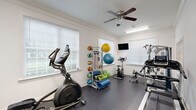 Located at 2118 Silvercrest Dr in Myrtle Beach, SC, this single-floor community facility is thoughtfully organized into distinct functional areas designed to serve residents' everyday needs. The primary fitness area is bright and welcoming, featuring white walls, crown molding, and durable dark rubber flooring. Multiple windows fitted with blinds allow natural light to fill the space, complemented by overhead fluorescent lighting and a ceiling fan that ensures comfortable air circulation during workouts. The gym is well-equipped with a range of cardio machines including a treadmill, elliptical trainer, stationary bikes, and a recumbent cycle. A multi-station cable strength machine provides comprehensive resistance training options, while a freestanding rack holds an assortment of medicine balls, resistance bands, stability balls, and exercise mats for versatile functional fitness. A wall-mounted flat-screen television adds entertainment during workouts. A secondary area, accessible through a glazed French door, offers additional space for cardio equipment. The facility also includes a dedicated laundry room, finished in the same clean white palette with concrete flooring. This room is equipped with multiple washers and dryers and features a window with a pleasant outdoor view, providing a practical and well-lit space for residents.