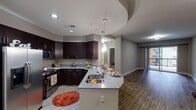 Located in the heart of downtown Raleigh, NC, this single-floor residence offers a well-designed open-concept layout with modern finishes throughout. The kitchen is a standout feature, equipped with stainless steel appliances including a French door refrigerator, microwave, and range. Rich dark cabinetry contrasts elegantly with granite countertops, while a curved peninsula with an undermount sink provides both additional workspace and casual seating. Stylish pendant lights illuminate the peninsula beneath a distinctive curved ceiling detail, adding architectural character to the space. Wood-look flooring extends throughout the main living areas, lending warmth and continuity. The living area is generously proportioned and bright, benefiting from natural light that pours in through sliding glass doors leading to a private balcony with outdoor views. The bedroom is carpeted for comfort and features a large window and direct access to a well-appointed bathroom. The bathroom includes a dark-cabinetry vanity with granite countertop and a walk-in glass-enclosed shower with tile surround. A spacious walk-in closet with wrap-around shelving and hanging rods rounds out the private quarters. The overall ambiance is contemporary and clean, with neutral grey tones providing a versatile, move-in-ready aesthetic.