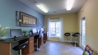This single-floor community business center is designed to offer a functional and comfortable environment for residents and visitors alike. The space is thoughtfully laid out, featuring a row of dedicated computer workstations built into a sleek, dark granite-topped counter with warm wood cabinetry below — providing ample workspace for multiple users simultaneously. Ergonomic adjustable bar-style chairs accompany each station, ensuring comfort during extended use. On the opposite side of the room, a high-top table paired with modern black bar stools offers an additional area for casual meetings or independent work. The interior is painted in a welcoming combination of cool blue and warm yellow tones, lending the space a bright and inviting atmosphere. A large window dressed with sheer white curtains allows generous natural light to fill the room, complementing the overhead fluorescent lighting. Decorative wall art adds a touch of personality, while potted greenery introduces a refreshing natural element. The tile flooring is clean and durable, well-suited to a high-traffic shared environment. Overall, the space strikes a practical balance between utility and aesthetic warmth, making it a versatile hub for work and connectivity.
