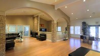 This single-story space showcases a striking open-concept design defined by soaring vaulted ceilings and elegantly curved archways that frame the living areas with architectural character. The most distinctive feature is the series of stacked stone columns, which anchor the space and serve as natural dividers between zones while adding a rich, textural contrast to the smooth, warm-toned walls.

Recessed lighting is integrated throughout the ceiling, complementing the generous natural light that filters in through multiple windows fitted with white shutters. The continuity of light-toned hardwood flooring across the space enhances the sense of openness and flow between areas.

A raised half-wall partition, clad in matching stacked stone, creates a subtle separation between the living and entertainment areas without disrupting the expansive feel of the interior. The entertainment nook is tucked neatly within this division, providing a defined media area.

The overall ambiance is warm and inviting, blending contemporary architectural details with natural materials. The combination of arched openings, stone accents, and open sightlines gives the space a refined yet comfortable character throughout.