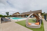 Located at 2523 Avenue M in Huntsville, Texas, this outdoor amenity complex offers a thoughtfully designed resort-style experience for its community. At the heart of the space is a large, shimmering swimming pool with a shallow entry area, surrounded by a generous concrete deck lined with lounge chairs and side tables for relaxed sunbathing. Distinctive wicker pod loungers with orange accent pillows are positioned poolside, providing stylish and comfortable retreats. A wooden pergola anchors an outdoor dining area furnished with tables and chairs, accented by string lights and stone pillar bases, creating an inviting space for gatherings. Grilling stations are conveniently located nearby. Turf lawn areas are scattered throughout, complemented by hanging egg chairs that add a playful, modern touch. A cornhole setup and additional open patio space encourage recreation and social activity. The complex is enclosed by sleek iron fencing, with natural greenery and open views of the surrounding landscape providing a refreshing backdrop. A clubhouse building with a pitched roof and wide covered porch faces the pool, serving as the architectural centerpiece of the space. The overall ambiance balances active outdoor living with comfortable relaxation in a well-appointed community setting.