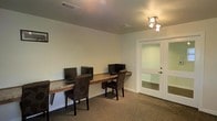 This single-floor space is designed with functionality and practicality in mind. The main area features a long, built-in granite or stone-topped desk that spans the width of the room, accommodating multiple computer workstations side by side — making it well-suited for a business center, study room, or collaborative workspace. Comfortable upholstered chairs in a dark, patterned fabric are positioned at each station, providing ergonomic seating for users.

The room is finished with neutral light-gray walls and carpeted flooring, creating a clean and professional atmosphere. Overhead, a multi-head track light fixture provides focused illumination, complementing the ceiling-mounted HVAC vents that ensure climate comfort throughout the space.

A standout architectural feature is the interior glass-panel door with a frosted finish, which leads to an adjoining room. This door allows natural light to pass between spaces while maintaining a degree of privacy. The adjoining room, partially visible through the glass, appears to be a similarly appointed area.

Overall, the space presents a well-organized, professional environment ideal for productivity-focused activities, with thoughtful design choices that balance utility and visual cleanliness.