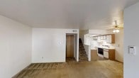 This two-story residence offers 2 bedrooms and 2.5 bathrooms within a well-utilized floor plan. The main level features an open living area finished with wall-to-wall carpet, neutral white walls, and a ceiling with a centrally placed HVAC vent for climate control. A wood-panel door near the base of the staircase provides secondary access or closet storage, adding practicality to the layout.

The kitchen is positioned adjacent to the living space in a semi-open configuration, separated by a partial dividing wall that creates a pass-through effect. It is equipped with light wood cabinetry, a white electric range, a white refrigerator, and a built-in microwave or vent hood above the stove. The kitchen flooring contrasts with the carpeted living area, offering an easy-to-clean surface suited for cooking and meal prep.

A carpeted staircase connects the main level to the upper floor, where the two bedrooms and additional bathrooms are located. The overall aesthetic is functional and straightforward, with a neutral color palette throughout. The layout makes efficient use of its footprint, providing essential living spaces across both levels.