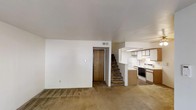 This two-story residence offers 2 bedrooms and 2.5 bathrooms within a well-utilized floor plan. The main level features an open living area finished with wall-to-wall carpet, neutral white walls, and a ceiling with a centrally placed HVAC vent for climate control. A wood-panel door near the base of the staircase provides secondary access or closet storage, adding practicality to the layout.

The kitchen is positioned adjacent to the living space in a semi-open configuration, separated by a partial dividing wall that creates a pass-through effect. It is equipped with light wood cabinetry, a white electric range, a white refrigerator, and a built-in microwave or vent hood above the stove. The kitchen flooring contrasts with the carpeted living area, offering an easy-to-clean surface suited for cooking and meal prep.

A carpeted staircase connects the main level to the upper floor, where the two bedrooms and additional bathrooms are located. The overall aesthetic is functional and straightforward, with a neutral color palette throughout. The layout makes efficient use of its footprint, providing essential living spaces across both levels.