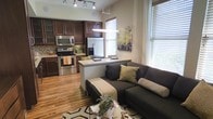 This single-level space showcases an open-concept layout that seamlessly connects the kitchen and living area, creating a cohesive and functional environment. The kitchen is well-appointed with rich, dark wood cabinetry featuring glass-front upper cabinets, a mosaic tile backsplash, and a full suite of stainless steel appliances, including a gas range, built-in microwave, and French door refrigerator. A peninsula with a light-colored countertop extends into the space, offering casual seating and serving as a natural dividing point between the kitchen and living area. Pendant lighting and a track lighting system illuminate the kitchen, while large windows dressed with horizontal blinds allow abundant natural light to fill the living space. The living area is anchored by a dark sectional sofa accessorized with patterned throw pillows, positioned over a geometric area rug atop warm hardwood flooring that runs throughout the entire space. Wall art adds a decorative touch, and the overall ambiance is modern, comfortable, and inviting.
