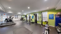 This fitness center is a spacious, single-floor facility designed to support a full range of workout routines. The interior is defined by a bold color scheme, with vibrant lime-green accent walls contrasting against clean white surfaces, creating an energizing atmosphere. Durable rubber flooring covers the entire space, providing both safety and comfort underfoot.

The strength training zone along the green-accented wall houses a variety of cable machines, selectorized weight stations, and multi-functional gym equipment, offering comprehensive options for resistance training. A well-stocked dumbbell rack with a flat bench provides additional free-weight capabilities, complemented by a stability ball and additional functional training accessories. A dedicated stretching and open floor area provides ample room for mat work and mobility exercises.

The cardio section features a row of Matrix-brand treadmills and elliptical machines positioned alongside a large floor-to-ceiling mirror, which enhances the sense of space and allows users to monitor their form. Recessed lighting throughout ensures bright, even illumination, while multiple windows invite natural light and offer views of the surrounding greenery. Framed fitness-themed artwork decorates the walls, and a water cooler station adds a practical convenience. The overall layout is organized and open, making the space accessible and motivating for users of all fitness levels.