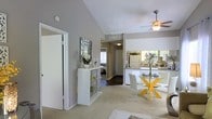 This single-story space is defined by its striking vaulted ceilings, which lend an airy, expansive quality to the interior. The open-concept layout connects the living, dining, and kitchen areas in a cohesive flow, making the space feel generously proportioned. Soft, neutral gray walls pair with plush carpeting in the main living areas, while dark hardwood-style flooring adds contrast in the transitional zones and hallway.

The living area benefits from a large sliding glass door dressed with vertical blinds, allowing ample natural light and providing an outdoor view. A ceiling fan with integrated lighting serves the dining area, which is positioned adjacent to the kitchen, separated by a breakfast bar counter. The kitchen features white cabinetry and full-size appliances.

A decorative display shelf and wall art add character along the hallway, which connects to the bathroom. The bathroom includes a wide vanity with white cabinetry, a well-lit mirror above a single sink, and hardwood-style flooring. The overall ambiance is clean, bright, and welcoming, with a consistent neutral palette throughout.