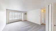 This efficiently designed 1-bedroom, 1-bathroom unit offers a functional single-floor layout across a compact but well-utilized footprint. The combined kitchen and living room serves as the heart of the unit, with the living area featuring neutral wall-to-wall carpeting, a large picture window dressed with vertical blinds that invites generous natural light, and a wall-mounted air conditioning unit alongside baseboard heating for year-round climate control. The kitchen is outfitted with warm wood-toned cabinetry — including both upper and lower cabinets — a white electric range, refrigerator, and a double-basin sink set into light countertops, all over durable tile-style flooring. The bedroom is a comfortable, well-proportioned room with soft carpeting, a window with outdoor views, baseboard heating, and direct access to a dedicated closet, providing practical storage. The full bathroom features tile flooring, a bathtub/shower combination with tiled surround, a toilet, and a window for natural ventilation. The unit's clean, light-toned palette throughout creates a bright and straightforward living environment suitable for those seeking a no-frills, functional residence.