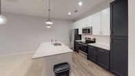 Located at 15928 White St in Charlotte, NC, this single-floor residence showcases a clean, modern design with neutral tones and quality finishes throughout. The open-concept layout flows seamlessly between living and dining areas, highlighted by plush carpeting, recessed lighting, and a striking globe pendant light fixture. Black-framed doors and windows add a bold contemporary contrast to the soft gray walls, while large windows invite natural light and outdoor views into the main living space.

The kitchen is a standout feature, offering a large central island topped with speckled granite countertops and an undermount sink with a professional-style faucet. Two pendant lights illuminate the island, complementing the recessed lighting above. Upper white shaker cabinets pair elegantly with dark lower cabinetry, and the suite of stainless steel appliances — including a French door refrigerator, electric range, and built-in microwave — rounds out the functional and stylish design. Wood-look flooring anchors the kitchen and extends into adjacent areas.

The bathroom is thoughtfully appointed with a tub/shower combination, curved curtain rod, granite-topped dark vanity, and wood-look flooring that ties the space together. An additional open room with recessed lighting and neutral carpeting offers flexible use as a bedroom or bonus space. Overall, the home presents a cohesive, move-in-ready aesthetic with modern upgrades throughout.