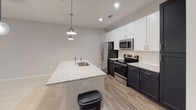 Located at 15928 White St in Charlotte, NC, this single-floor residence showcases a clean, modern design with neutral tones and quality finishes throughout. The open-concept layout flows seamlessly between living and dining areas, highlighted by plush carpeting, recessed lighting, and a striking globe pendant light fixture. Black-framed doors and windows add a bold contemporary contrast to the soft gray walls, while large windows invite natural light and outdoor views into the main living space.

The kitchen is a standout feature, offering a large central island topped with speckled granite countertops and an undermount sink with a professional-style faucet. Two pendant lights illuminate the island, complementing the recessed lighting above. Upper white shaker cabinets pair elegantly with dark lower cabinetry, and the suite of stainless steel appliances — including a French door refrigerator, electric range, and built-in microwave — rounds out the functional and stylish design. Wood-look flooring anchors the kitchen and extends into adjacent areas.

The bathroom is thoughtfully appointed with a tub/shower combination, curved curtain rod, granite-topped dark vanity, and wood-look flooring that ties the space together. An additional open room with recessed lighting and neutral carpeting offers flexible use as a bedroom or bonus space. Overall, the home presents a cohesive, move-in-ready aesthetic with modern upgrades throughout.