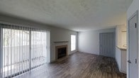 This single-floor space centers around a welcoming living area that combines practical design with comfortable features. The room is anchored by a fireplace with a simple mantel and tile surround, serving as a natural focal point. Dark wood-style laminate flooring runs throughout, adding warmth and contrast against the light gray walls. A textured ceiling adds subtle visual interest overhead.

Natural light enters generously through a large sliding glass door fitted with vertical blinds, offering outdoor access and views of the exterior. An additional window near the fireplace further brightens the space. The neutral gray wall tones create a versatile backdrop suitable for a variety of interior styles.

A gray interior door along the far wall provides access to other areas of the home, reinforcing the functional layout of the space. The open floor plan allows for flexible furniture arrangement, making the living area adaptable to different needs. Overall, the space presents a clean, move-in ready environment with a straightforward aesthetic, well-suited for everyday living.