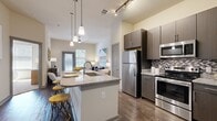 This thoughtfully designed 2-bedroom, 1-bathroom residence features a single-floor open-concept layout that maximizes both functionality and style. The kitchen serves as the centerpiece of the living area, boasting dark espresso cabinetry, granite countertops, a mosaic tile backsplash, and a full suite of stainless steel appliances including a refrigerator, range, and built-in microwave. A peninsula island with pendant lighting overhead provides additional prep space and seating, flowing seamlessly into the living area. Rich hardwood-style flooring runs throughout the main living spaces, complemented by track lighting and soaring ceilings that enhance the sense of openness. The living area benefits from large windows that invite generous natural light. Both bedrooms are carpeted and well-sized, with the spacious primary bedroom offering ample room for a full furniture arrangement. The second bedroom also features large windows for natural light. A dedicated walk-in closet with wire shelving provides organized storage. The bathroom is finished with a dark vanity, granite countertop, wood-style flooring, and a tiled shower-tub combination. The overall ambiance is modern and clean, with a neutral color palette that creates a bright, welcoming atmosphere throughout.