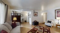 This single-floor residence offers a thoughtfully designed open-concept layout that seamlessly connects the living, dining, and kitchen areas. The living room is a comfortable and inviting space, featuring warm hardwood flooring, clean white walls, and soft ambient lighting provided by wall sconces and a floor lamp. A bold geometric area rug in rich red and earth tones anchors the seating area, which includes a plush sofa and a classic armchair. A dark cube ottoman serves as a functional centerpiece, complemented by framed wall art that adds a touch of character to the space. A decorative folding screen panel provides a stylish visual division between the living and dining areas. The dining area accommodates a bistro-style table with ornate metal chairs, accented by a distinctive circular wall decoration that enhances the room's aesthetic. Adjacent to the dining area, a passthrough hallway leads to the kitchen, where white appliances are visible. A hallway extending from the living area provides access to the private rooms beyond, with doors visible in the background. The overall ambiance is bright, clean, and welcoming, with a cohesive neutral palette punctuated by warm decorative accents throughout.