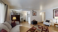 This single-floor residence offers a thoughtfully designed open-concept layout that seamlessly connects the living, dining, and kitchen areas. The living room is a comfortable and inviting space, featuring warm hardwood flooring, clean white walls, and soft ambient lighting provided by wall sconces and a floor lamp. A bold geometric area rug in rich red and earth tones anchors the seating area, which includes a plush sofa and a classic armchair. A dark cube ottoman serves as a functional centerpiece, complemented by framed wall art that adds a touch of character to the space. A decorative folding screen panel provides a stylish visual division between the living and dining areas. The dining area accommodates a bistro-style table with ornate metal chairs, accented by a distinctive circular wall decoration that enhances the room's aesthetic. Adjacent to the dining area, a passthrough hallway leads to the kitchen, where white appliances are visible. A hallway extending from the living area provides access to the private rooms beyond, with doors visible in the background. The overall ambiance is bright, clean, and welcoming, with a cohesive neutral palette punctuated by warm decorative accents throughout.