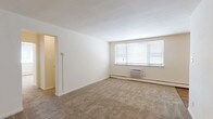 This single-floor residential unit offers a practical and well-organized layout across several distinct rooms. The spacious living area is finished with wall-to-wall beige carpeting and neutral cream-toned walls, creating a clean and versatile backdrop. A wall-mounted air conditioning unit and baseboard heating provide year-round climate control, while a large multi-pane window allows ample natural light to fill the room. An adjacent area, visible from the living space, flows seamlessly toward the kitchen and entry door.

The kitchen features warm honey-toned wood cabinetry with ample upper and lower storage, a gas range, a full-size refrigerator, and laminate countertops. The flooring transitions to hardwood-style planks in this area, adding a practical and attractive contrast to the carpeted living spaces. A pass-through counter connects the kitchen to the main living area.

The bedroom is generously proportioned, with continued carpeting, a closet, baseboard heating, and a window providing natural light. The overall layout, as seen in the dollhouse view, is compact yet efficient.

The bathroom is a standout feature, showcasing retro charm with pink ceramic wall tiles accented by teal border detailing, a matching teal soaking tub, and a classic basket-weave tile floor. A window above the tub brings in natural light, completing this distinctive vintage-inspired space.