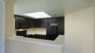 This single-floor space showcases a well-appointed kitchen as its central feature. The kitchen is defined by a striking contrast between rich, dark espresso-toned cabinetry and light-colored countertops with subtle speckled detailing. Upper and lower cabinets provide generous storage, complemented by black appliances, including a range, built-in microwave, and refrigerator, creating a cohesive, modern aesthetic. A peninsula counter extends outward, offering additional workspace and a natural division between the kitchen and the adjoining area. A prominent rectangular skylight is positioned above the kitchen, drawing in natural light and brightening the space from above. The ceiling is finished in a smooth, neutral tone, and the walls are painted in a clean, light beige, contributing to an open and airy atmosphere. HVAC vents are discreetly integrated into the ceiling, ensuring functional comfort throughout. The overall design balances bold, dark finishes with lighter surrounding elements, resulting in a kitchen space that feels both purposeful and visually dynamic.