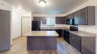 This two-story home offers a practical and comfortable layout across both levels. The main floor features a spacious kitchen outfitted with dark espresso cabinetry, light-toned countertops, a full suite of black appliances including a refrigerator, gas range, over-the-range microwave, and dishwasher. A generously sized center island provides additional prep and storage space, while wood-look flooring and a window over the sink add functionality and natural light. Adjacent to the kitchen, the main living area is finished with neutral carpet and features a ceiling fan with light fixture, creating a relaxed atmosphere. A carpeted staircase connects the main level to the upper floor. The upper level hosts multiple bedrooms, each finished with soft carpet and windows that allow in natural light and exterior views of the surrounding neighborhood. At least one bedroom features vaulted ceilings, adding a sense of height and openness. The home includes multiple full bathrooms, each equipped with a tub and shower combination set against clean white surrounds and light-toned tile flooring. Throughout the home, the neutral color palette and consistent finishes create a cohesive, move-in ready environment.
