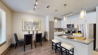 This stylish single-floor residence combines contemporary design with thoughtful functionality across its well-defined living spaces. The open-concept kitchen is a standout feature, boasting white shaker cabinetry, granite countertops, a subway tile backsplash, and stainless steel appliances including a range and microwave. A peninsula island with bar seating and elegant pendant lighting creates a natural gathering point, while track lighting illuminates the space efficiently. Wood-look plank flooring flows seamlessly through the kitchen and into the adjacent dining area, which offers ample room for a full dining set beneath a striking framed art piece. The living room is bright and inviting, with large windows that allow generous natural light and views of the surrounding exterior. A console with framed wall art adds a polished, curated touch to the entry area. The bedroom is carpeted for comfort and features a window with exterior views, providing a calm and restful atmosphere, with direct access to the en-suite bathroom. The bathroom is finished with large-format tile flooring, a dual-sink granite vanity with white cabinetry, framed mirrors, and a spacious glass-enclosed walk-in shower clad in stone-toned tile — a refined and functional retreat.