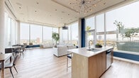 Located at 1140 Wellington St W in Ottawa's vibrant Hintonburg neighbourhood, this contemporary amenity space is designed to impress. The open-concept layout is anchored by soaring floor-to-ceiling windows that flood the interior with natural light and frame sweeping panoramic views of the Ottawa skyline. Light-toned hardwood flooring runs throughout, complementing a clean, modern aesthetic accented by bold navy blue walls and architectural columns.

The lounge area offers generous seating with plush, neutral-toned sofas arranged to take full advantage of the expansive city views. A striking sputnik chandelier serves as a focal point overhead, adding a mid-century modern design element. A ceiling-mounted projector enhances the space's functionality for gatherings and presentations.

The fully equipped kitchen features light wood cabinetry, white tile backsplash, quartz countertops, stainless steel appliances, and a large island with bar seating — ideal for entertaining. A bright dining area with floor-to-ceiling glazing overlooks the outdoor terrace.

The rooftop terrace extends the living space outdoors, offering a wood-deck patio with pergola shade structures, lounge seating, umbrella-shaded tables, a barbecue station, and lush greenery — all set against breathtaking elevated views of Ottawa.