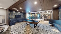 Located at 1 Midtown Bridge Approach in Hackensack, NJ, this single-floor amenity space is designed with a contemporary aesthetic that blends sophisticated materials and bold design choices throughout. The entrance lobby features soaring double-height ceilings, exposed brick columns, and light hardwood flooring, with floor-to-ceiling glass walls framing views of an outdoor courtyard. A vibrant geometric-patterned area rug and curved lounge seating establish a welcoming tone from the moment you enter.

The lounge and game area is a standout zone, anchored by a full-size pool table set atop a textured area rug. A dramatic curved sectional sofa in slate blue faces a large flat-screen TV mounted on a dark accent wall adorned with striking gold geometric line work. Warm wood-paneled ceilings with recessed lighting and exposed beam detailing add depth and visual interest overhead.

Adjacent to the game lounge, a demonstration kitchen offers a generous island with bar-height seating in teal velvet chairs, wood cabinetry, open shelving, and an industrial-style pot rack suspended from the ceiling. A curated gallery wall and a bar-style seating counter complete the social atmosphere. Together, these areas create a cohesive, upscale communal environment designed for gathering and entertainment.