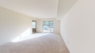 This single-floor unit is designed with a clean, neutral aesthetic throughout, featuring cream-toned walls and light-colored carpet in the main living and sleeping areas. The spacious living room is a standout feature, boasting elevated tray ceilings that add architectural dimension and a sliding glass door that opens to a balcony, flooding the space with natural light and providing outdoor views of surrounding greenery.

An arched passage connects the living area to an additional room, which also benefits from direct balcony access via another sliding glass door, making it a versatile space for various uses. The kitchen is equipped with rich cherry wood cabinetry, a gas range with overhead ventilation, a refrigerator, and light-toned countertops, all set on tile flooring that extends into the adjoining entry area.

The unit includes two bathrooms: a full bath featuring a tiled tub-shower combination, a wood-framed vanity mirror, and tile flooring, and a second bathroom with a matching cherry wood vanity and medicine cabinet mirror. The bedrooms offer generous proportions, multiple windows with blinds for natural light and privacy, and closet storage. The overall layout is functional and well-organized, with a cohesive design language maintained across all rooms.