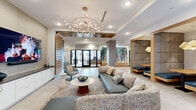 Located at 4 Tuscan Blvd in Salem, New Hampshire, this single-floor amenity space offers a thoughtfully designed environment that blends contemporary aesthetics with functional comfort. The welcoming lobby lounge serves as the heart of the space, anchored by a sculptural branch-like chandelier and furnished with a generous sectional sofa, accent chairs, and a large wall-mounted television set against a slatted wood feature wall. Light-toned tile flooring flows throughout, unifying the various zones seamlessly. An archway framed in warm wood slat detailing leads to a secondary lounge area centered on a dramatic floor-to-ceiling marble fireplace wall with gold veining, flanked by curved upholstered armchairs. A dedicated café-style dining area features bistro-height tables, mixed upholstered seating, warm pendant lighting, and a wood-paneled ceiling, with natural light entering through large windows. A cozy semi-private booth nook, lined with textured padded walls and topped with a sculptural pendant light, provides an intimate setting for meetings or casual conversation. Curated artwork is displayed throughout the corridors and common areas, adding cultural depth to the space. A fitness center, accessible via glass-paneled doors, completes the amenity offering. The overall design is modern, warm, and polished.