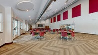 Located at 17833 Whitney Rd in Strongsville, Ohio, this single-floor multipurpose space offers a generously sized, open-plan room designed for group gatherings, dining, and community activities. The architectural design draws immediate attention with its soaring ceilings, which vary in height across the room, creating a dynamic and open atmosphere. Exposed cylindrical ductwork runs along the ceiling, lending an industrial-modern aesthetic that is complemented by circular recessed lighting fixtures and recessed downlights throughout.

One of the room's most striking design elements is the series of large, bold crimson acoustic panels mounted high along the white walls, providing both visual impact and sound management. The floor features an eye-catching herringbone-patterned carpet with alternating neutral tan and deep burgundy tones, adding texture and visual interest to the expansive floor area.

Natural light filters in generously through multiple large windows and glass exit doors along the perimeter, offering views of an adjacent outdoor patio area. The room is furnished with round tables and colorful upholstered chairs in red and green patterns, arranged to accommodate flexible seating configurations. A wall-mounted flat-screen television and a recessed accent nook with deep crimson walls further enhance the space's functionality and character.
