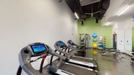 This fitness center offers a comprehensive workout environment designed to support a wide range of exercise routines. The space features a sleek, modern aesthetic with polished concrete floors, exposed black structural ceilings, and a combination of crisp white and energizing green accent walls that create a motivating atmosphere throughout.

The cardio zone is generously appointed with a row of commercial-grade treadmills, each equipped with individual touchscreen displays, as well as elliptical trainers and a stationary bike — providing ample options for aerobic exercise. Large floor-to-ceiling windows and a glass entry door flood this area with natural light while offering views to the exterior.

The strength and functional training area is set against the vibrant green accent wall and features a multi-functional cable machine, adjustable weight benches, and free weights. A well-organized rack displays an assortment of stability balls, resistance bands, medicine balls, and yoga mats, making functional and flexibility training highly accessible.

Overhead linear LED lighting and ceiling fans ensure the space remains well-lit and comfortable during use. The open floor plan allows for easy movement between zones, while the thoughtful layout accommodates both individual training and small group workouts. Overall, the fitness center delivers a professional, fully-equipped environment suited for a broad spectrum of fitness goals.