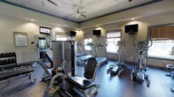 This fitness center is thoughtfully designed to provide a comprehensive workout environment on a single floor. The space is outfitted with a full suite of exercise equipment, including multiple treadmills, elliptical trainers, and strength training machines, offering users a wide range of workout options. Each cardio machine is paired with its own dedicated wall-mounted monitor, allowing users to stay entertained during their sessions. Additional wall-mounted flat-screen TVs are positioned throughout the room for shared viewing. The space benefits from several windows fitted with horizontal blinds, allowing natural light to filter in while maintaining privacy. Ceiling fans overhead, complemented by recessed lighting, ensure the room remains well-lit and comfortable during use. The flooring features a durable, dark-toned surface suited for high-traffic athletic use. Crown molding with a contrasting accent color adds a refined architectural detail along the ceiling perimeter, elevating the overall aesthetic of the room. The warm, neutral-toned walls contribute to an energizing yet comfortable atmosphere, making this fitness center a functional and inviting space for daily exercise.