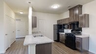 This 2-bedroom, 2-bathroom residence is designed for comfortable, modern living across a single, well-organized floor. The open-concept kitchen and living area serves as the heart of the home, featuring rich wood-toned cabinetry, granite-style countertops, and a central peninsula island with a built-in sink and pendant lighting overhead. Black appliances — including a gas range, refrigerator, microwave, and dishwasher — provide a bold contrast against the lighter countertops. Durable wood-style flooring runs throughout the main living areas, complementing the neutral, light-toned walls. The living room is bright and airy, benefiting from natural light through a large window and a private exterior door, with a ceiling fan adding comfort overhead. The primary bedroom is generously sized, offering plush carpeting, a ceiling fan, and direct access to a well-equipped en-suite bathroom. The primary bathroom features a soaking tub with shower, a granite-style vanity with dark cabinetry, and a walk-in closet with built-in shelving. The second bedroom also features carpet flooring, a ceiling fan, and a large window with outdoor views. A dedicated closet provides additional storage within the unit.