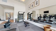 Located at 6146 Liberty Hall Dr in Leland, NC, this single-floor fitness and wellness facility is thoughtfully designed to support a comprehensive approach to health and exercise. The entry corridor makes a strong first impression with its deep charcoal accent walls, herringbone-patterned wood-look tile flooring, and geometric lantern pendant lighting that guides visitors through the space. The main fitness area features soaring ceilings with a distinctive wood-paneled tray ceiling detail and an abundance of natural light streaming through large windows and glass doors. The gym is outfitted with a full row of TRUE brand cardio equipment — including treadmills, ellipticals, and a recumbent bike — alongside a comprehensive strength training zone with a multi-use rack, TRX suspension systems, and a complete dumbbell rack. Full-wall mirrors amplify the sense of space throughout. Adjacent to the main gym, a dedicated yoga and therapy studio offers light wood plank flooring, floor-to-ceiling mirrors, a built-in storage platform, and a wall-mounted television, creating a serene environment for group classes and personal wellness practices. Framed coastal artwork adds a cohesive, calming aesthetic throughout the entire facility.